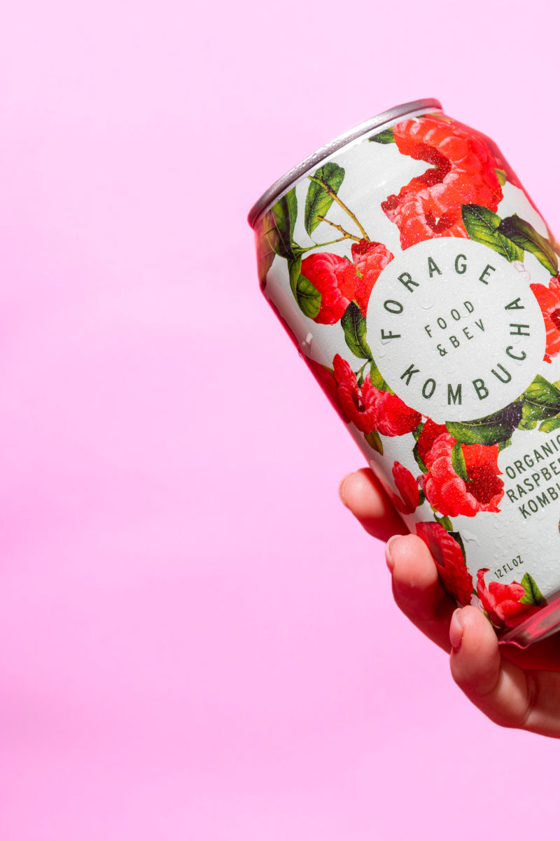 A can of Forage Kombucha held in front of a pink backdrop.