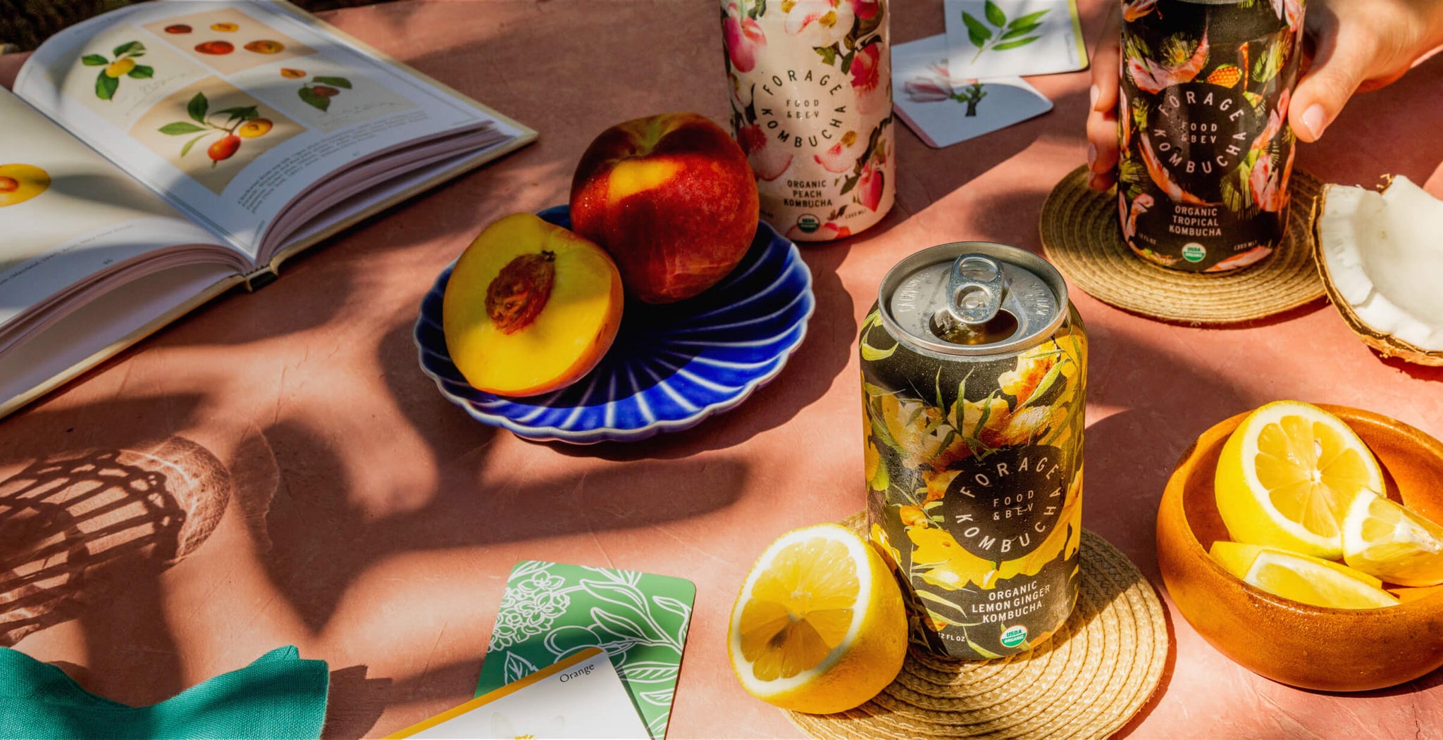 A tables cape with an open book, peaches, lemons, playing cards, and some cans of Forage Kombucha.