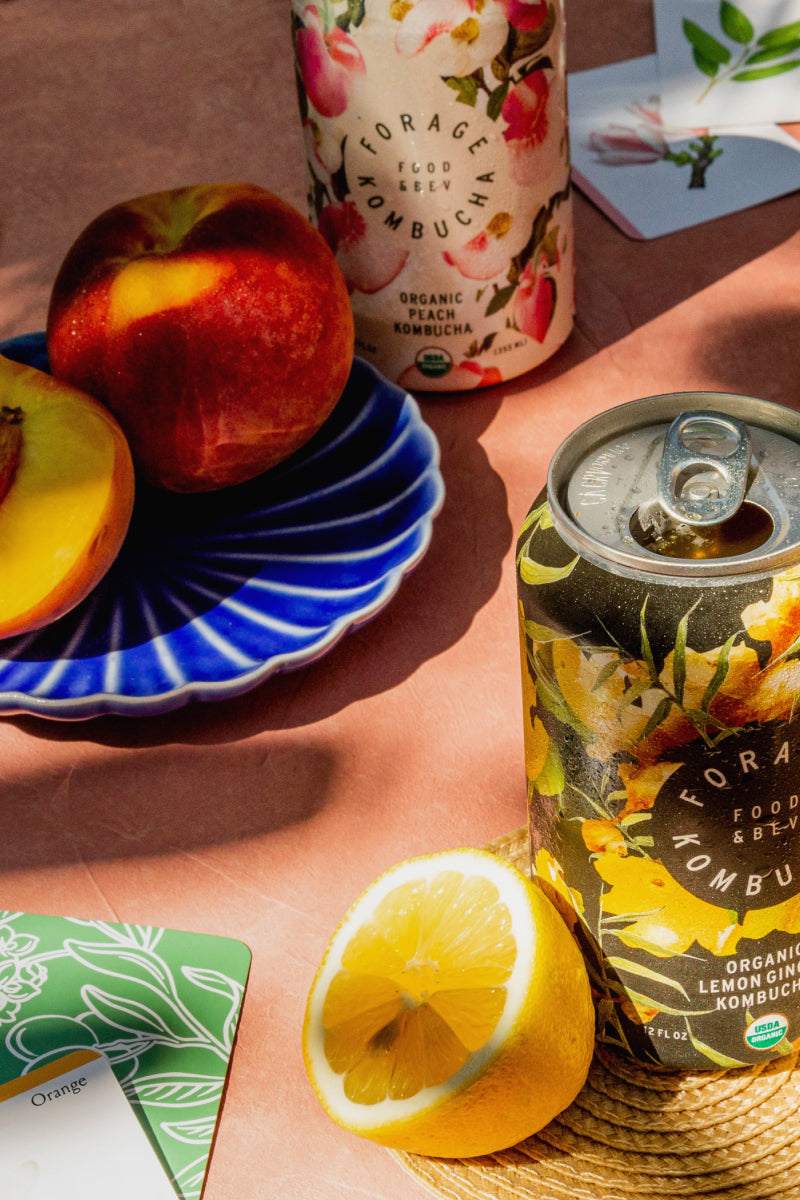 A tables cape with an open book, peaches, lemons, playing cards, and some cans of Forage Kombucha.