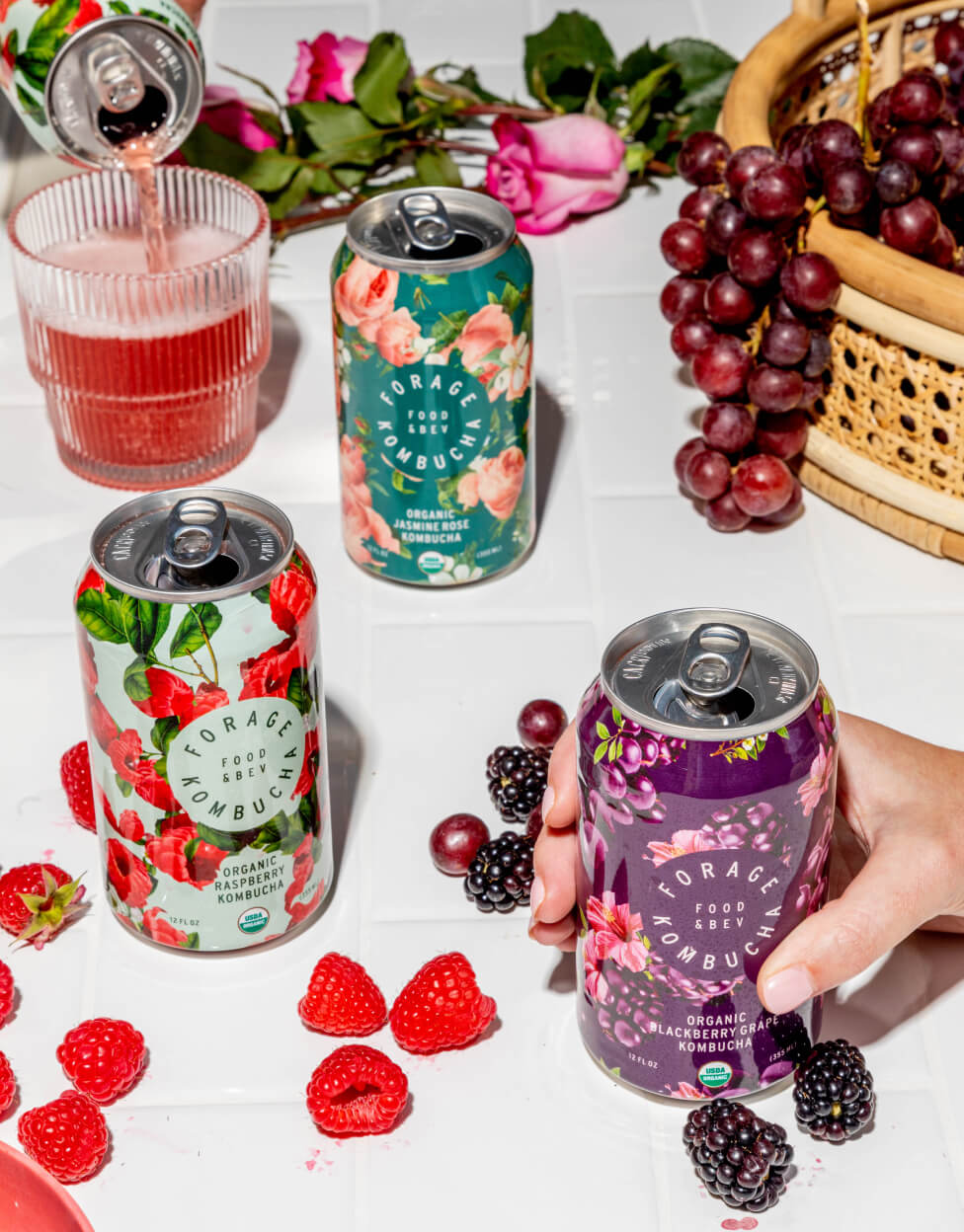 A variety of Forage Kombucha cans set on a white tablecloth surrounded by raspberries, blackberries, and grapes.